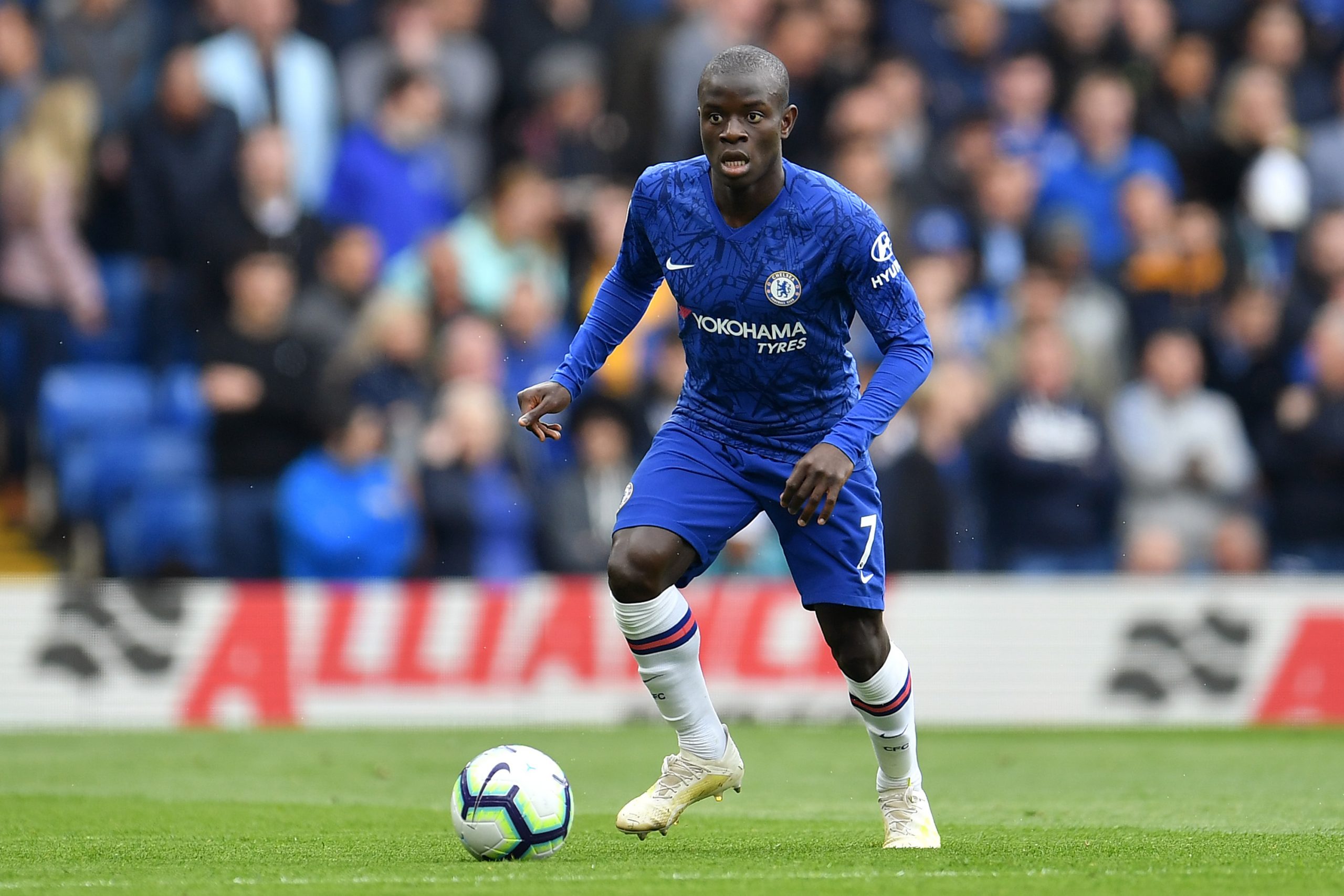  N'Golo Kante is playing soccer for Chelsea, wearing a blue jersey with white sleeves and blue shorts, with the ball at his feet.